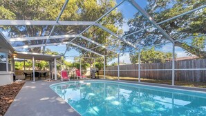 A covered swimming pool with a patio area, chairs, and a barbecue grill.