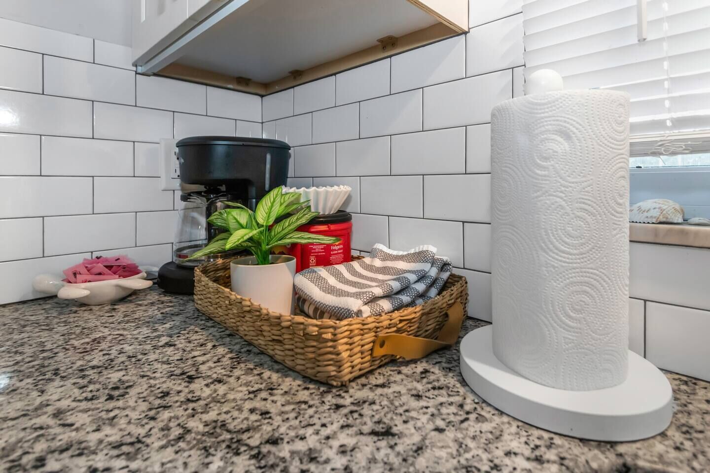 Una cocina con paredes de azulejos blancos, una encimera de granito, una cafetera, una cesta con una planta, un recipiente rojo y un rollo de toallas de papel.