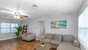 A spacious living room with hardwood flooring, a ceiling fan, a large window with blinds, a painting of a beach, and a potted plant.