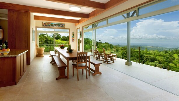 A spacious dining area with a large table and chairs, a kitchen with wooden cabinets, and large windows offering a view of lush greenery and a distant landscape. - Arenal-Area Jungle Lodge — Pool Access, 5BR/6.5BA, Sleeps 20 (Cabanga)