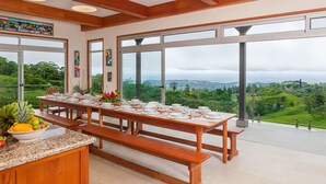A dining area with a large table set for a meal, large windows offering a view of a lush landscape, and wooden beams on the ceiling. - Arenal-Area Jungle Lodge — Pool Access, 5BR/6.5BA, Sleeps 20 (Cabanga)