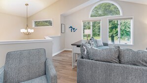 A spacious living room with a high ceiling, a dining area with a table and chairs, and large windows offering a view of greenery. - Beautiful home, quiet neighborhood w hot tub. Close to shopping, beach & trails. (Holland)