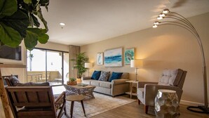 A living room with a sofa, armchairs, a coffee table, and a floor lamp. There are paintings on the wall and a plant in the corner.