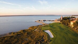 Luftaufnahme eines Golfplatzes mit einem Gewässer, einem Leuchtturm und Gebäuden im Hintergrund.