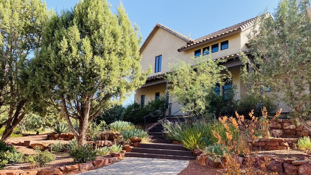 A house with a paved driveway leading to a set of stairs, surrounded by various plants and trees.