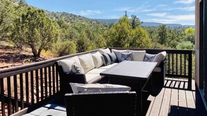 An outdoor deck with a sofa and table, surrounded by a wooden railing, overlooking a hilly landscape.