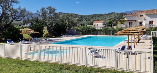 Piscina rectangular con cerca blanca, rodeada de sillas de descanso y sombrillas, con casas y colinas al fondo.