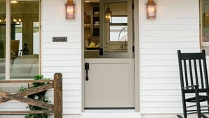 A white building with a grey door, two lantern-style lights, and a small porch area with a chair.
