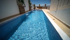 Una piscina con un diseño de agua azul cristalina, rodeada por un muro blanco y un suelo color beige, con vista al cielo y un edificio lejano.
