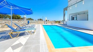 Una piscina con sillas de descanso azules y una sombrilla azul, un edificio blanco y un muro de piedra al fondo.