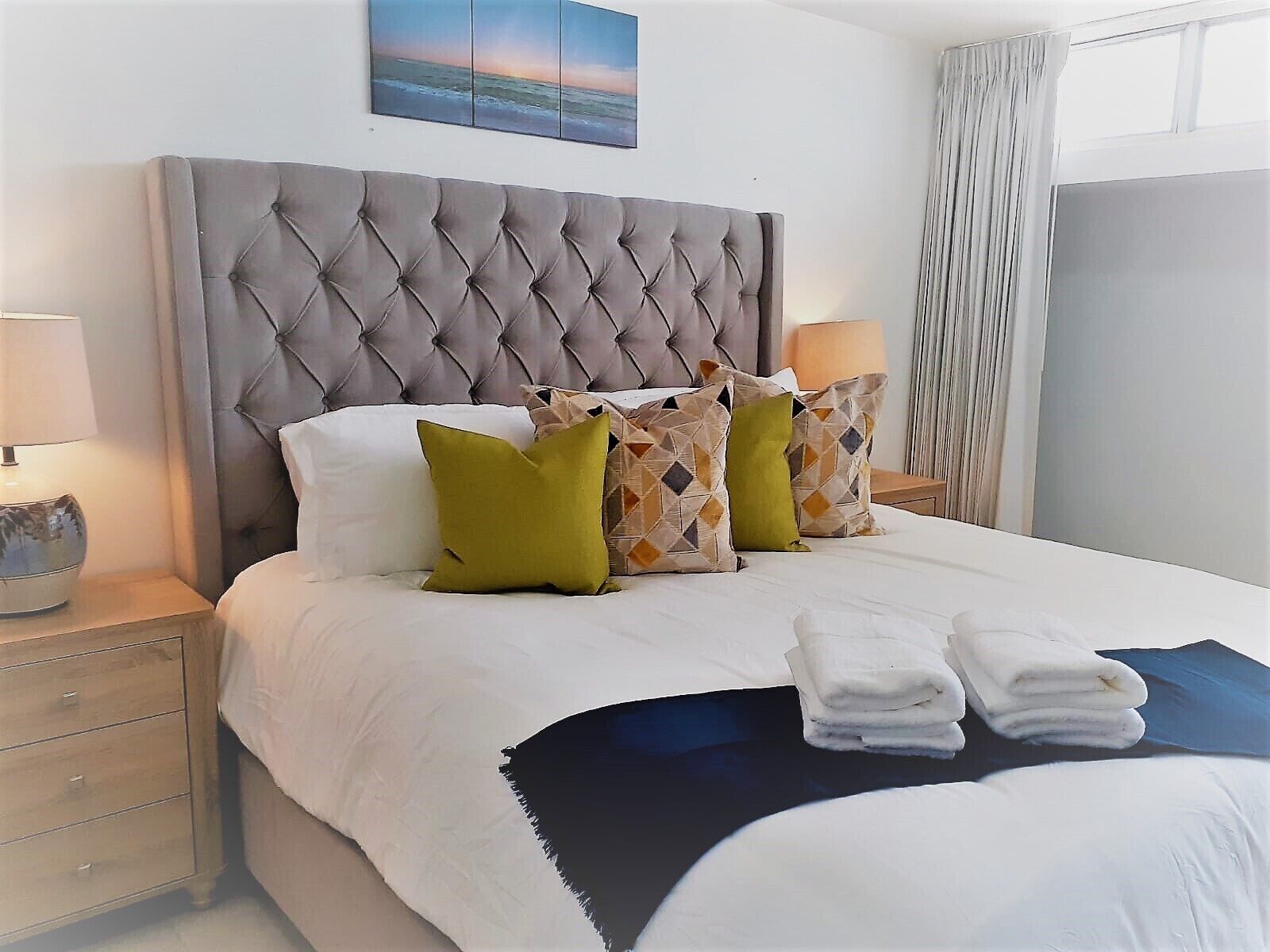 Un lit bien fait, doté d’une tête de lit capitonnée, de draps blancs et de coussins décoratifs. On y trouve deux tables de chevet avec des lampes et un petit tiroir.