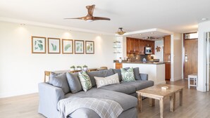 A modern living room with a grey sectional sofa, a wooden coffee table, and a ceiling fan. There is a kitchen area with wooden cabinets and a microwave.