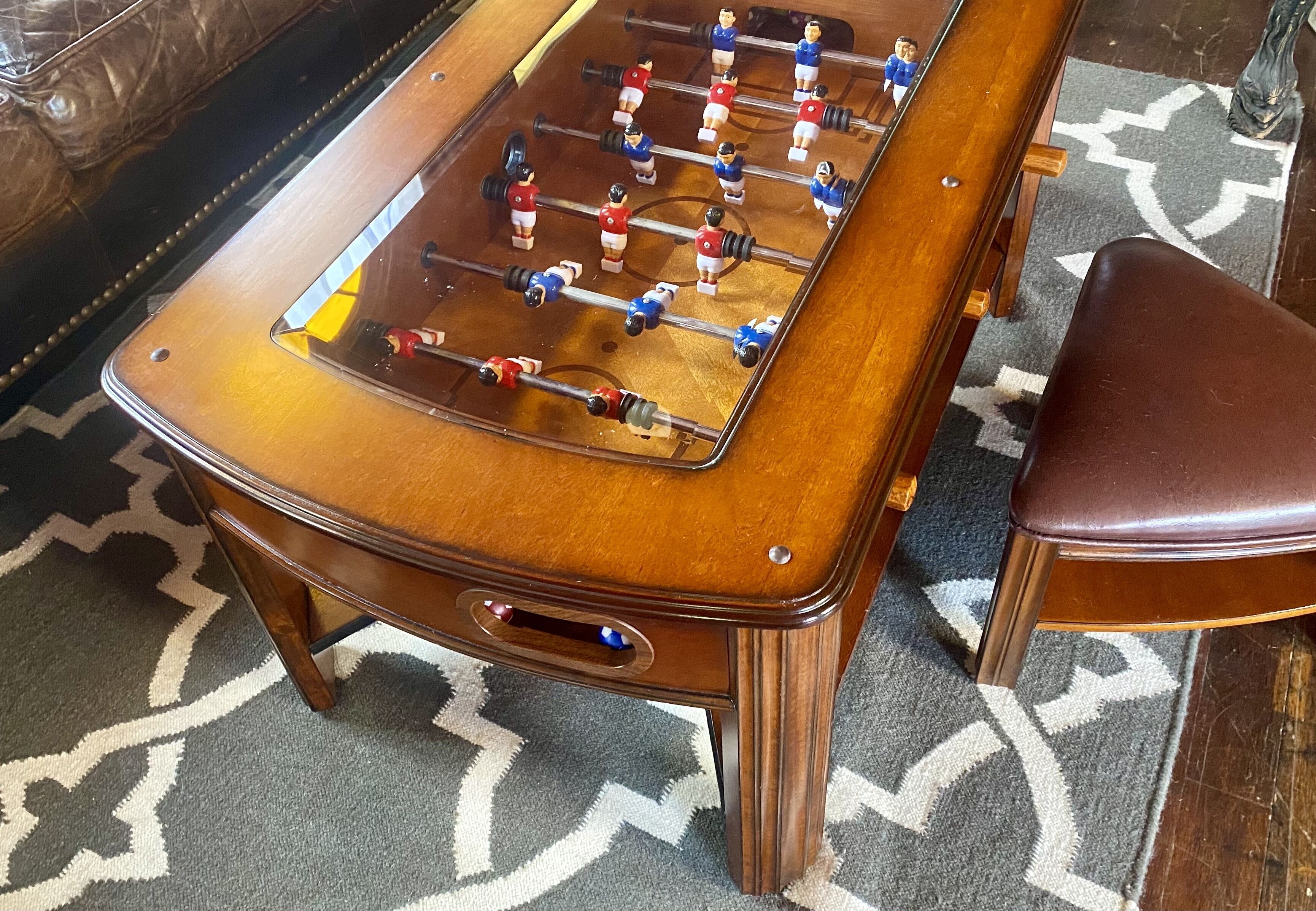 A wooden foosball table with a clear top, positioned on a patterned carpet.