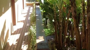 Una terraza de madera que lleva a una casa con un jardín, destacando árboles de plátano y abundante vegetación.