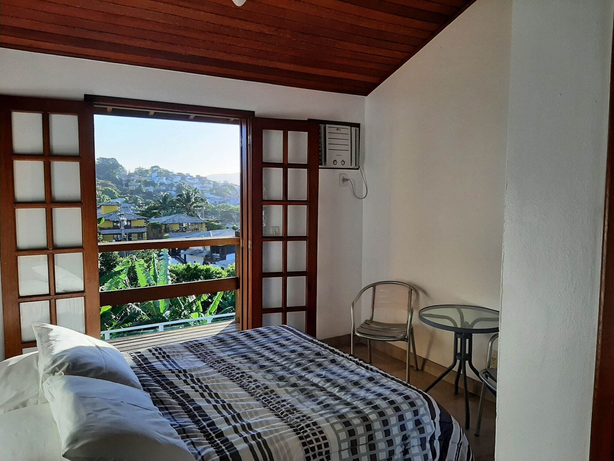 Une chambre à coucher avec un lit, une petite table et une chaise. Il y a une fenêtre qui donne sur des immeubles et de la verdure.