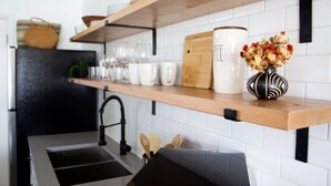 A kitchen with white tiled walls, a black refrigerator, a wooden shelf with white mugs, a white pitcher, and a vase with flowers.