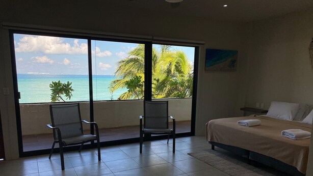 Ein Hotelzimmer mit einer großen Schiebeglastür, die auf einen Balkon mit Blick auf den Ozean und Palmen führt. Zwei Stühle und ein Bett mit beiger Tagesdecke und weißen Kissen sind vorhanden.