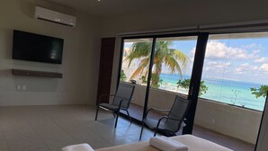 Ein Hotelzimmer mit Bett, zwei Stühlen, einem Fernseher und einem Balkon mit Blick auf das Meer und Palmen.