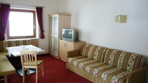 A small, furnished room with a sofa, a television on a cabinet, a dining table with chairs, and a window with curtains. - Alpenheim Joergele (Soelden)