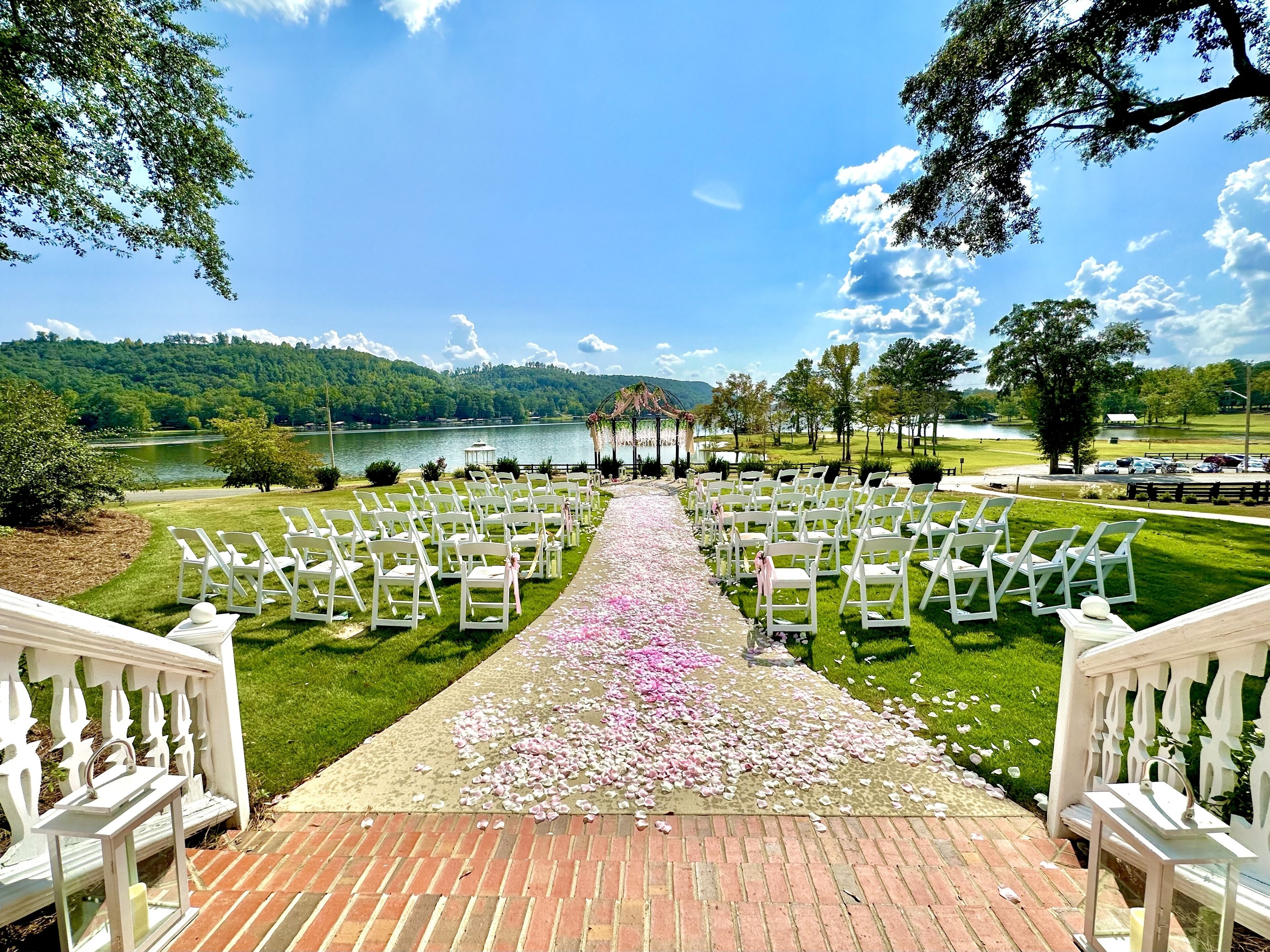Une allée de cérémonie de mariage avec des chaises blanches et un chemin de briques menant à un belvédère au bord d’un lac.