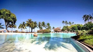 Una piscina cristalina con borde curvo, rodeada de palmeras y sombrillas de playa.