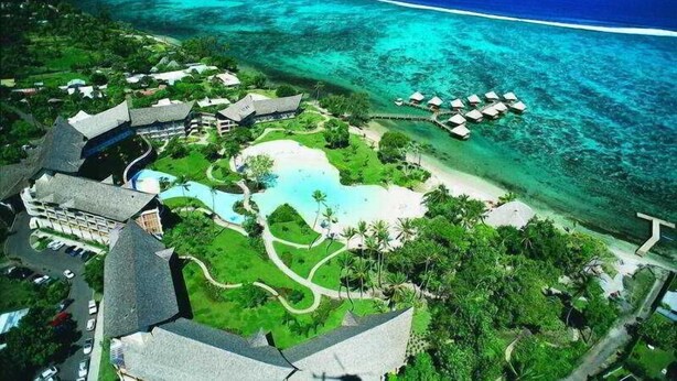 Vista aérea de un resort con una piscina grande, villas frente a la playa y bungalows sobre el agua.