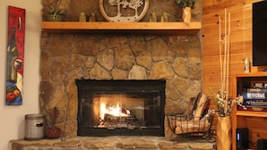 A stone fireplace with a fire, a wooden shelf above it, and a television on the wall to the right. - Lakefront Gem, Mtn views, dock + HOT TUB (Blairsville)