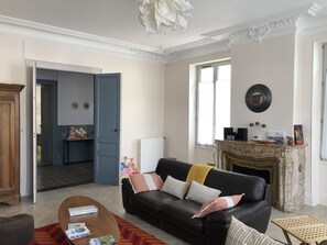 Ein Wohnzimmer mit einem schwarzen Ledersofa, einem Holztisch und einem Kamin. Eine Tür führt in einen anderen Raum, ein Fenster bietet einen Ausblick.
