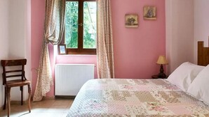 A bedroom with a bed, a chair, a window with curtains, a radiator, and a small table with a lamp. - Triantafillies Hotel (Volos)