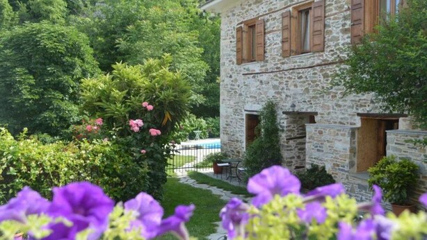 A stone building with wooden shutters, surrounded by lush greenery and a small pool. - Triantafillies Hotel (Volos)