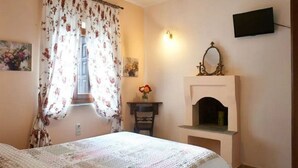 A bedroom with a bed, a window with floral curtains, a wall-mounted TV, a mirror above a fireplace, and a small table with a vase of flowers. - Triantafillies Hotel (Volos)