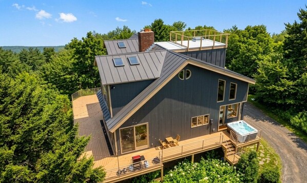 A two-story house with a deck, a hot tub, and a view of the forest. - Vermont`s Mountain Villa: Hot tub, Ski views, Pet+ (Andover)