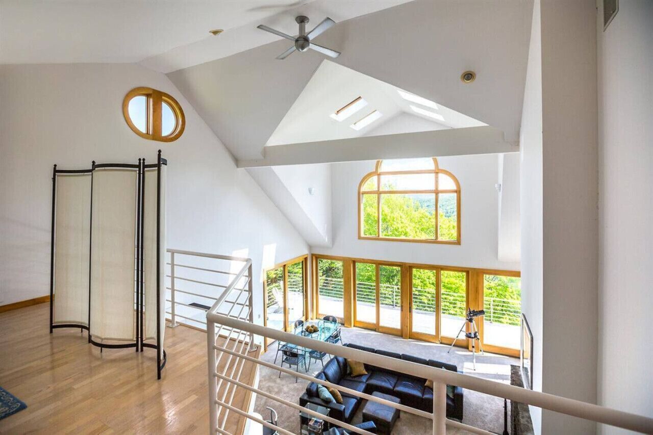 A spacious room with a high ceiling, a ceiling fan, a large window with a view of greenery, a black leather sofa, and a small table with chairs.