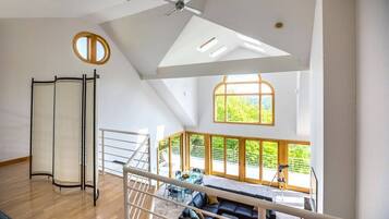 A spacious room with a high ceiling, a ceiling fan, a large window with a view of greenery, a black leather sofa, and a small table with chairs.