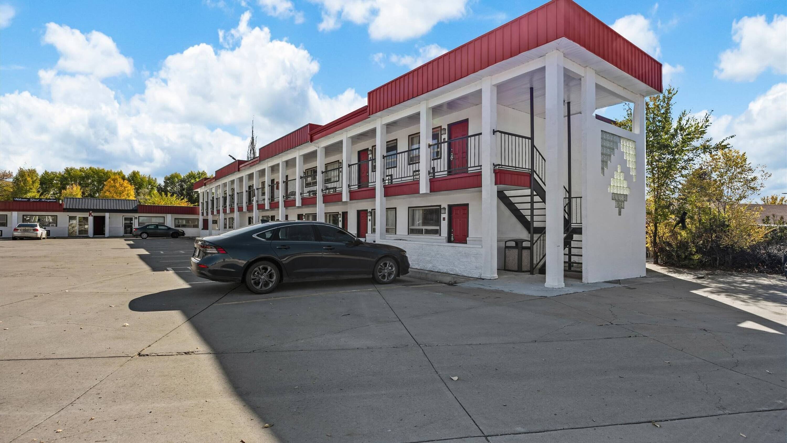 A multi-story motel with a red roof and white exterior, featuring a parking lot and a staircase leading to the entrance.