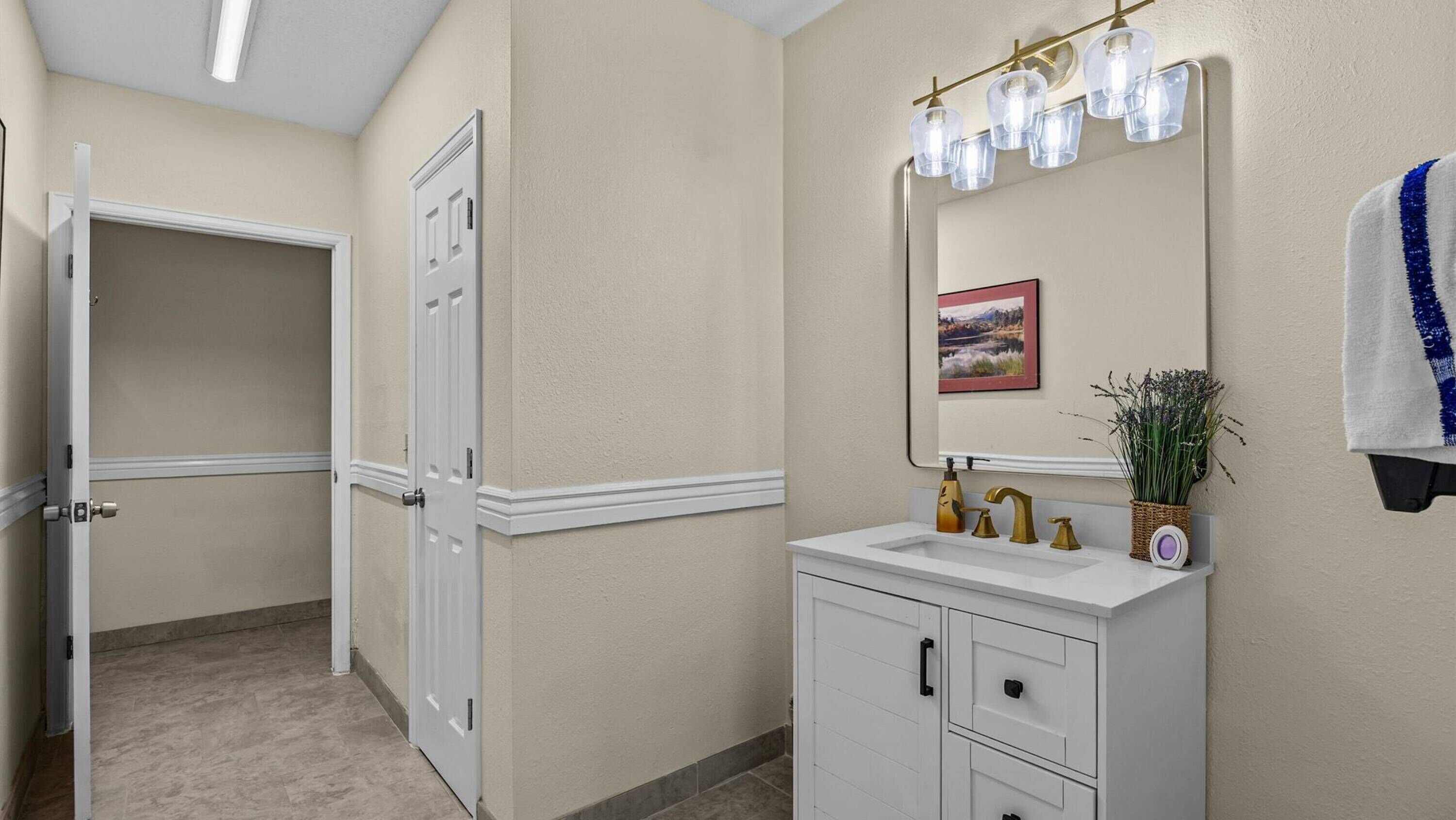 A bathroom with a white vanity, a mirror with lights, a framed picture, and a towel hanging on the wall.
