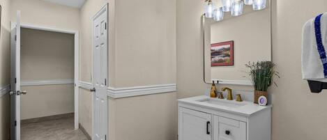 A bathroom with a white vanity, a mirror with lights, a framed picture, and a towel hanging on the wall.