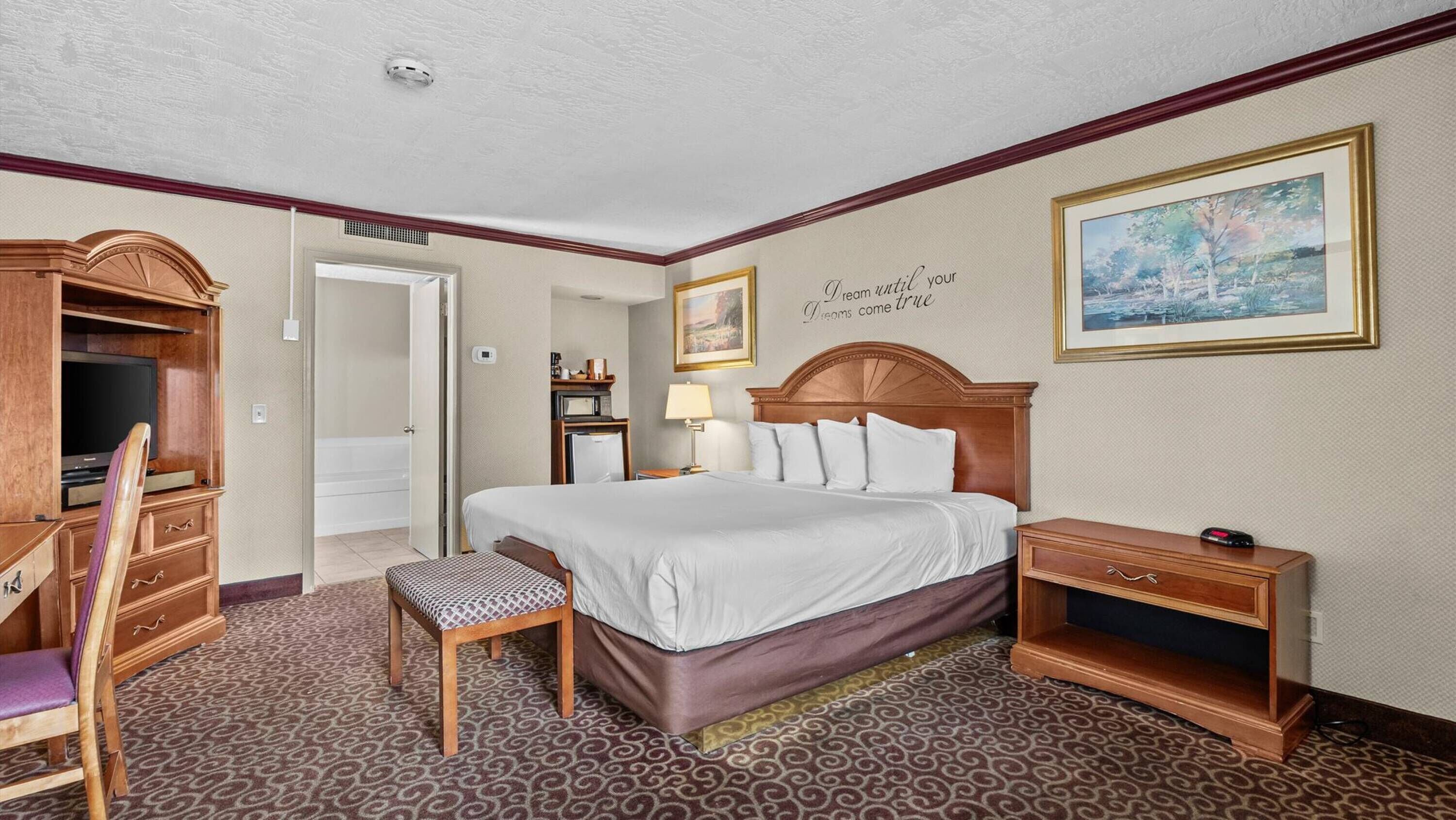 A hotel room with a large bed, a desk with a computer, a chair, a small table, a lamp, and a painting on the wall.