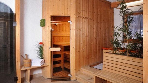A wooden sauna room with a bench, a bench with a towel, a small table with a towel and a plant, and a wooden door. - Appartementhaus Alpina (Bad Hofgastein)