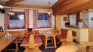A rustic wooden interior with a dining area featuring benches and a table, a kitchen area with a microwave, and a window with curtains. - Appartementhaus Alpina (Bad Hofgastein)