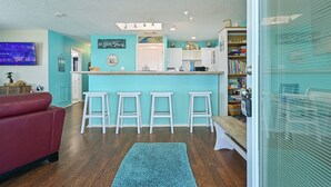 A kitchen with a teal accent wall, wooden flooring, a red couch, and a bar area with stools. - Sea Shed-Big yard in Jamaica Beach-Pet Friendly! (Jamaica Beach)
