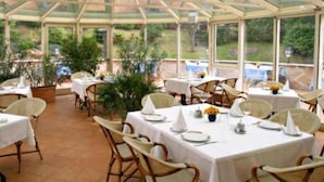A covered outdoor dining area with tables and chairs, large windows, and potted plants. - Mediterran Hotel (Budapest)