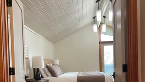 A bedroom with a sloped ceiling, a bed with a white comforter and a brown throw blanket, a nightstand with a lamp, and a wooden floor. - Spacious South Hill View Home, Private Mountain Setting & Hot Tub (Spokane)
