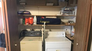 A compact laundry area with two washing machines, a dryer, and a shelf with various items including a Graco baby seat.