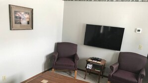 A living room with two purple armchairs, a wooden coffee table, a television, and a small side table with books and a dice.
