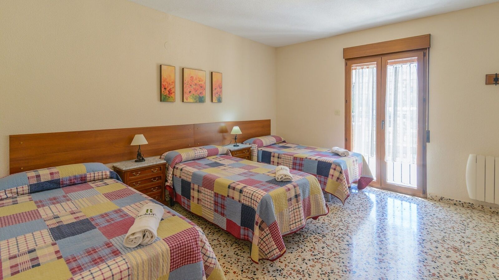 A hotel room with three beds, each with a quilted bedspread, a wooden headboard, bedside tables with lamps, and a large window with curtains.