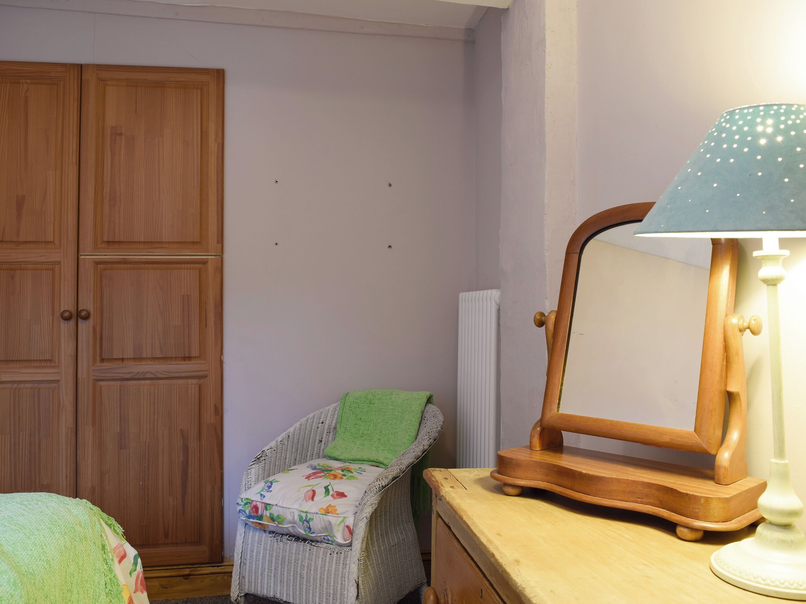 A room with a wooden wardrobe, a wicker chair with a floral cushion, a wooden side table with a lamp, and a radiator.
