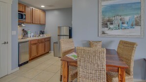 A kitchen with wooden cabinets, a refrigerator, and a dining area with wicker chairs and a wooden table. - Origin 1309: Gulf + Sunset Views + Ice Maker! Free Fun! (Panama City Beach)