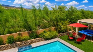 A swimming pool with a stone wall, a patio with red chairs and a red umbrella, and a view of a green landscape with trees and mountains in the background.