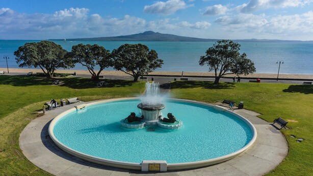 Ein runder Pool mit einer zentralen Fontäne, umgeben von einem gepflasterten Weg, Bäumen und Blick auf das Meer sowie einen Berg im Hintergrund.